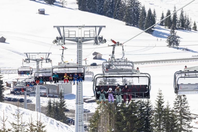 Sessellift mit Skifahrern fährt über verschneite Hänge, vorbei an Tannen und vereinzelten Hütten in Berglandschaft.