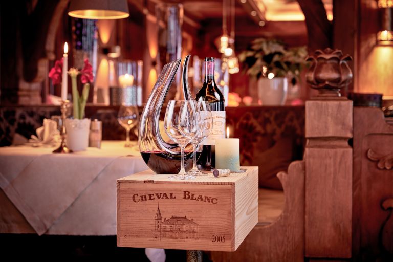 Holzkiste mit Aufdruck "CHEVAL BLANC 2005" trägt Weinflasche, Dekanter und Gläser in einem warm beleuchteten Restaurant.
