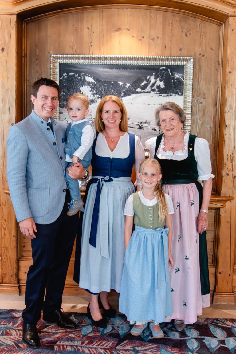 Fünfköpfige Familie posiert lächelnd in Trachten vor einer Holzwand mit gerahmtem Schwarzweiß-Bergfoto dahinter.
