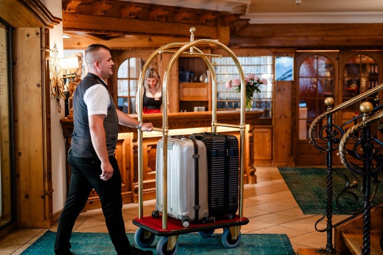 Gepäckwagen wird von einem Hotelangestellten mit zwei Koffern durch eine warme Holzhallen-Lobby zum Empfang geschoben.