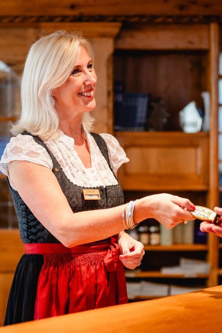 Frau gibt Schlüssel lächelnd über Holztresen und trägt traditionelle Tracht mit einem Namensschild.