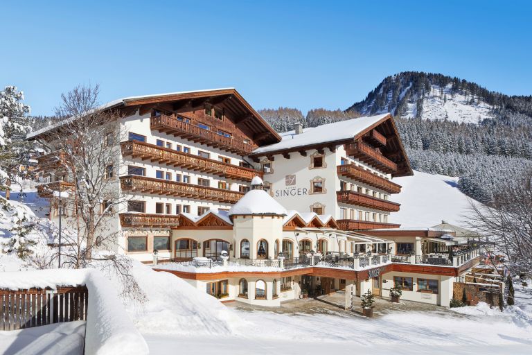 Alpenhotel "SINGER" steht verschneit, mit Holzbalkonen bedeckt, vor einem sonnigen, schneebedeckten Berghang.
