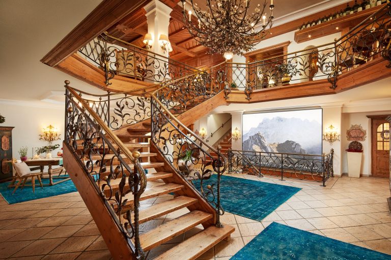 Geschwungene Holztreppe steigt empor, schmiedeeiserne Geländer schmücken sie, in einem hellen, alpenhaften Hotel-Foyer.