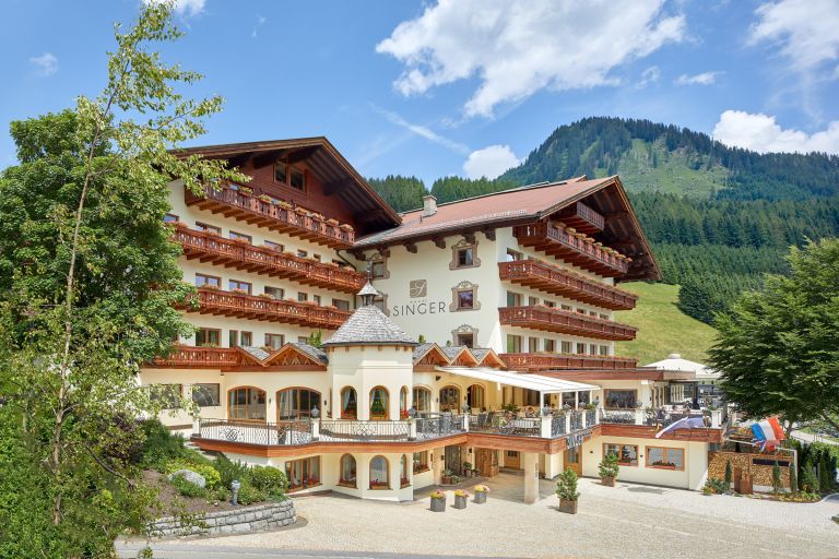 Hotel "SINGER" steht am Waldrand, zeigt hölzerne Balkone und eine große Terrasse vor grünen Alpenhängen und blauem Himmel.