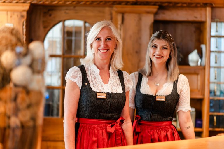 Zwei lächelnde Frauen in Dirndl und Namensschild stehen hinter einer Holztheke in einem rustikalen Gastraum.