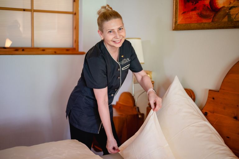 Hotelangestellte richtet lächelnd weiß bezogene Kissen auf einem Holzbett in einem hellen Gästezimmer. Auf der Uniform steht 'SINGER'.