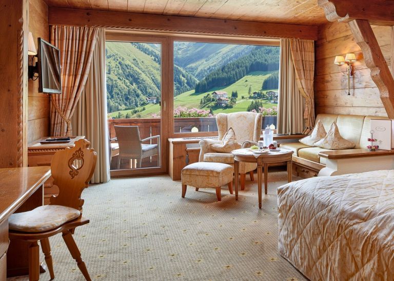 Beiger Sessel mit Fußhocker steht vor einer Balkontür mit Blick auf grüne Berge; Holzinterieur, Bett und Sofa im Zimmer.