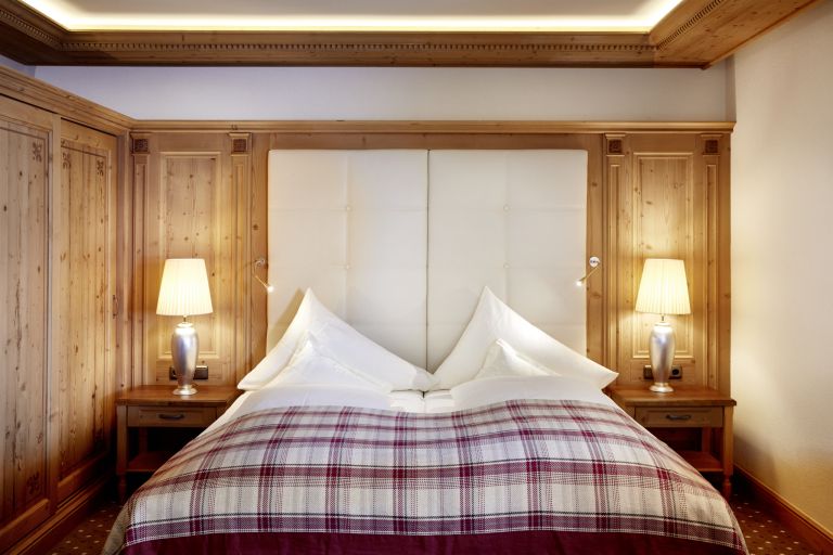 Doppelbett mit karierter Tagesdecke und weißen Kissen; eingerahmt von Holzpaneelen und zwei eingeschalteten Nachttischlampen.