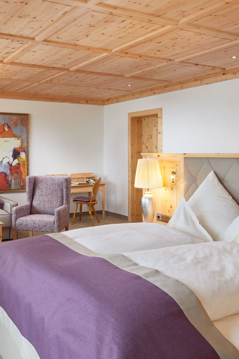 Ordentlich bezogenes Doppelbett mit grauem Kopfteil und Lampe daneben in einem Zimmer mit Holzdecke, Sessel und Schreibtisch.