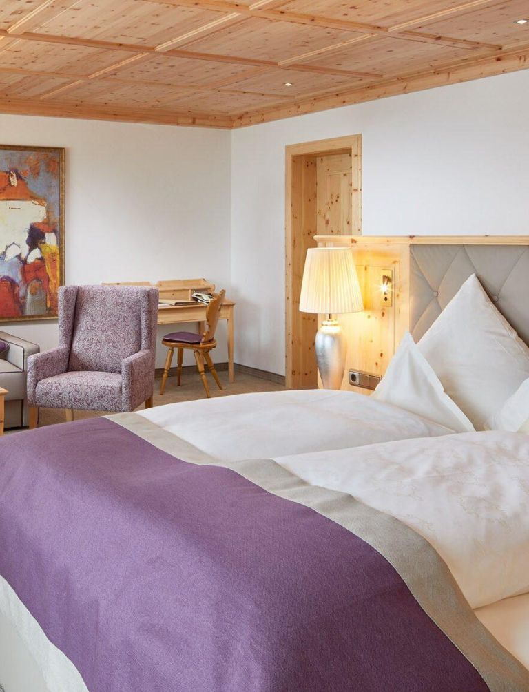 Het bed, opgemaakt met wit beddengoed en een paarse sprei, staat in een lichte kamer met een fauteuil, een bureau en een lamp.