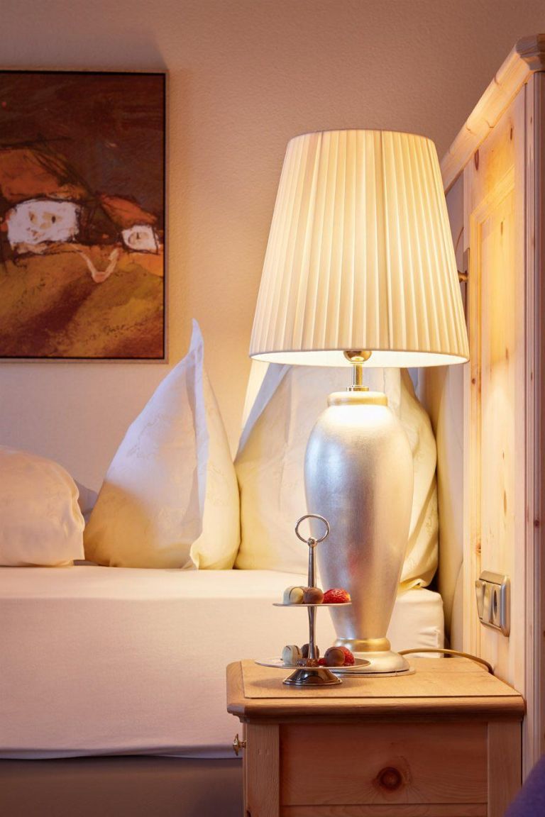 Nachttischlampe und Etagère mit Pralinen auf einer Holzkommode neben einem weiß bezogenen Bett; An der Wand ein Bild in Brauntönen.