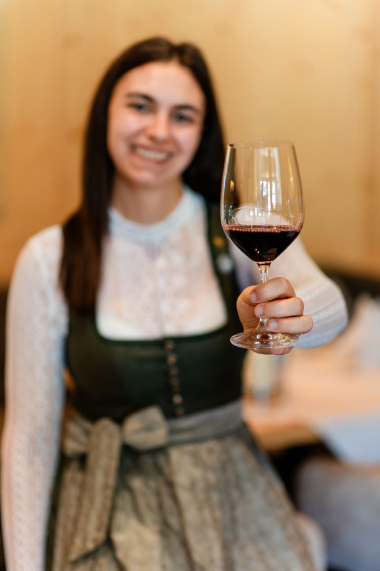 Weinglas mit Rotwein, gehalten von einer lächelnden jungen Frau im Dirndl, vor warmem Holzhintergrund in Innenraum.