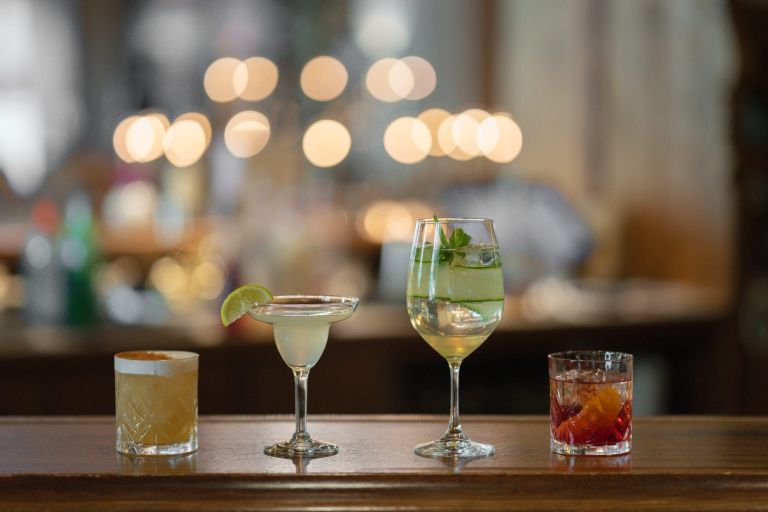 Vier Cocktails stehen auf einer Bar, mit Limette, Gurke und Orangenzeste garniert; unscharfe Lichter im Hintergrund.