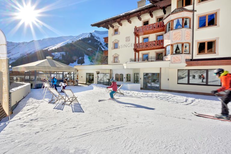 Kind fährt Ski vor Hotelterrasse und schneebedeckten Bergen im Hintergrund bei Sonnenschein. Auf der Fassade steht "SINGER".