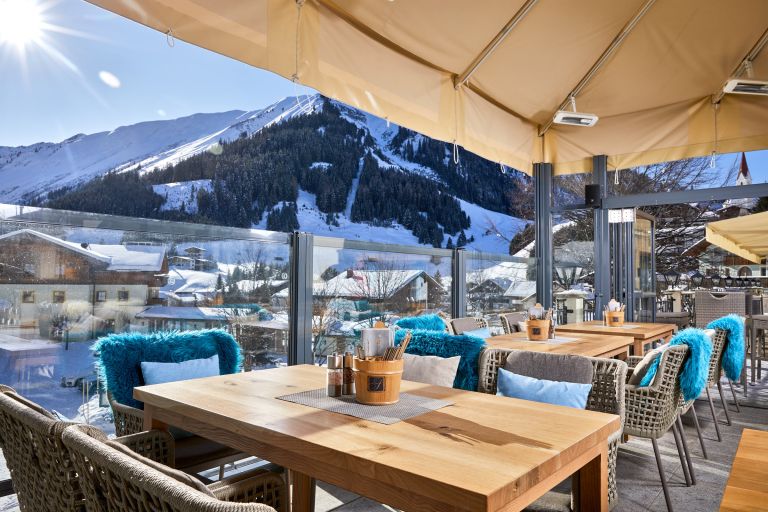 Terrassentische und Stühle mit blauen Fellen gedeckt, vor Glasbrüstung und schneebedeckten Bergen.