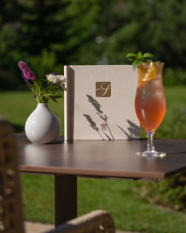 Leinengebundenes Menü steht auf einem Gartentisch und links daneben eine Vase mit Wildblumen und rechts ein Cocktailglas.