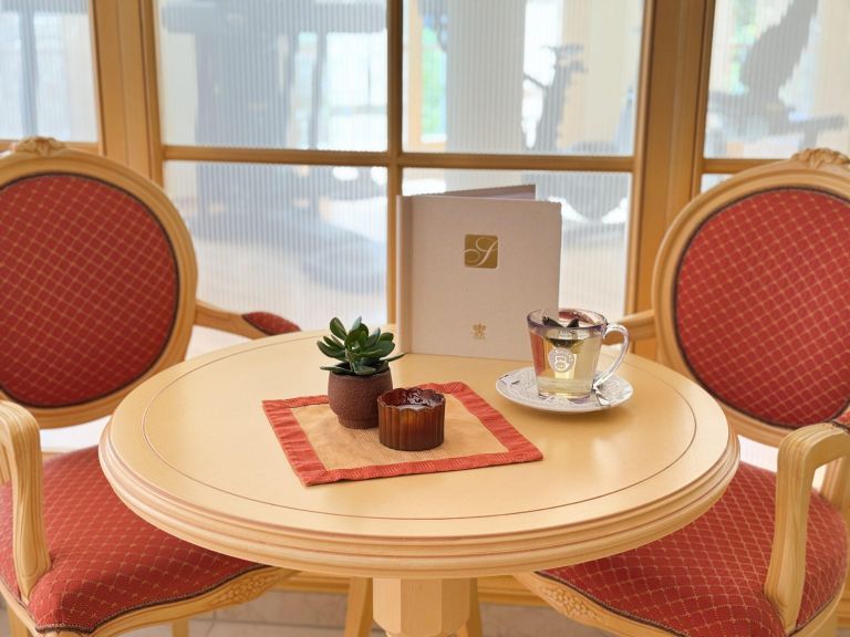 Runder Holztisch trägt Tasse mit Untertasse, Sukkulente und Menü; dahinter zwei rote Stühle und eine Fensterfront.