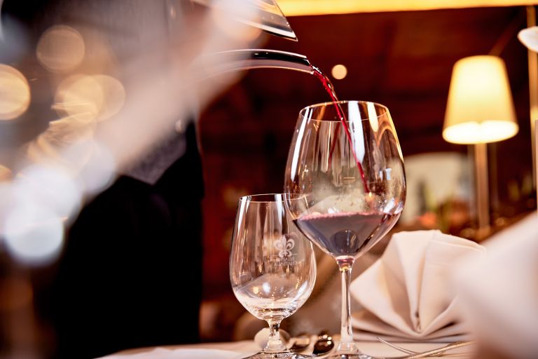 Großes Weinglas wird aus einer Karaffe mit Rotwein gefüllt, neben leerem Glas auf gedecktem Tisch im warmen Restaurantlicht.
