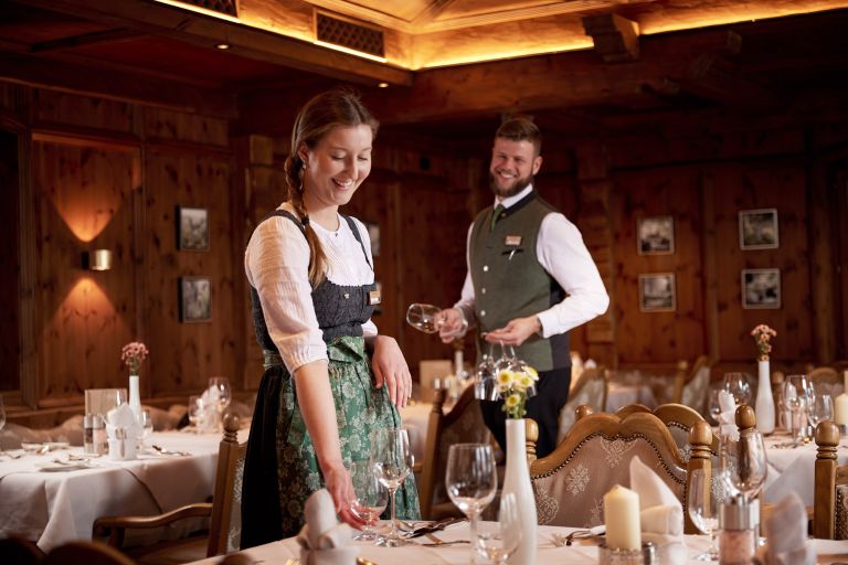Kellnerin stellt Weinglas auf Tisch und ein Kellner hält ein weiteres Glas in rustikalem, holzgetäfelten Speisesaal.