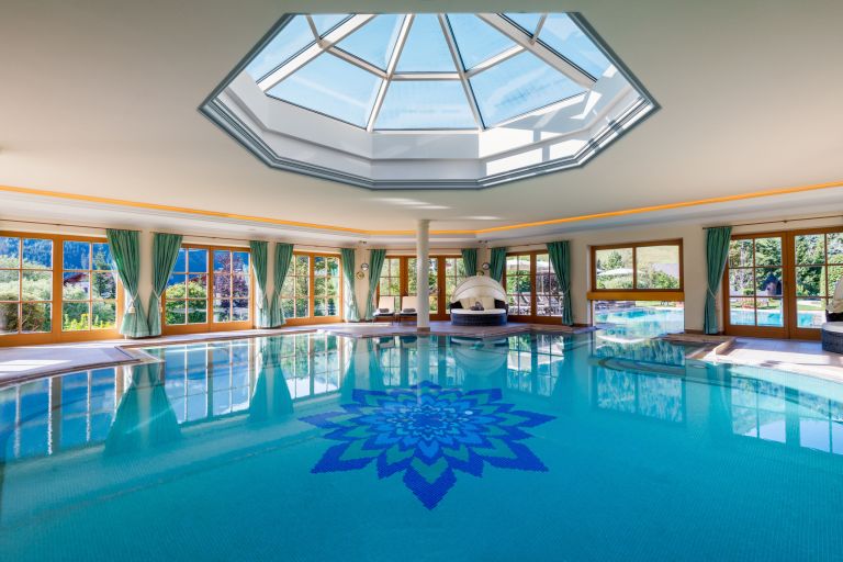 Innenpool mit blauem Blütenmosaik s umgeben von Panoramafenstern, grünen Vorhängen und Liegen.
