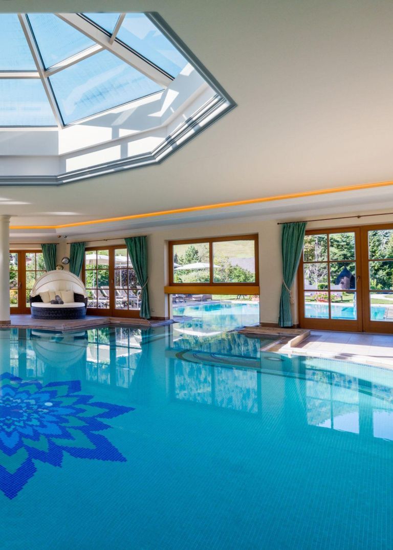 Schwimmbecken spiegelt Tageslicht aus einer Glaskuppel; angrenzende Fenster, grüne Vorhänge und Blick in den Garten.