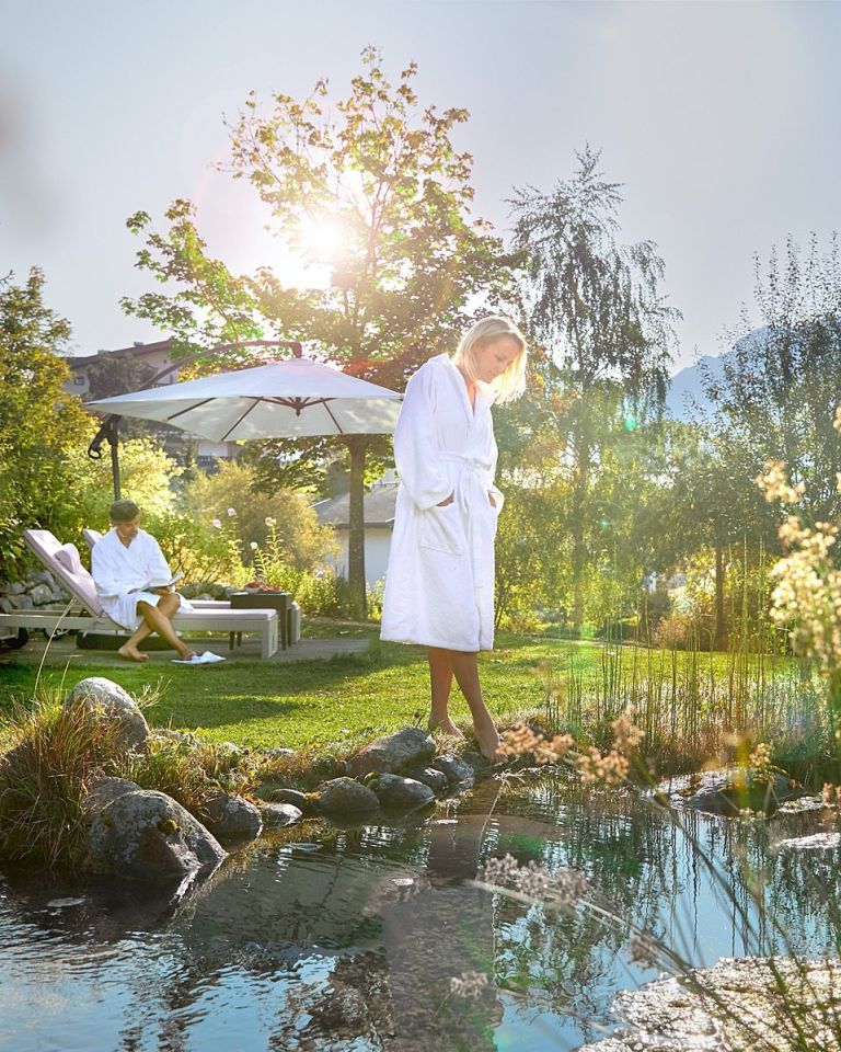 Frau im Bademantel steht am Teichrand und taucht Zehen ins Wasser; im Garten liest ein Mann unter einem Sonnenschirm.