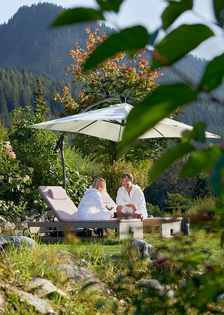 Paar sitzt auf Liegen in Bademänteln unter Sonnenschirm und entspannt im Garten vor bewaldeten Bergen.