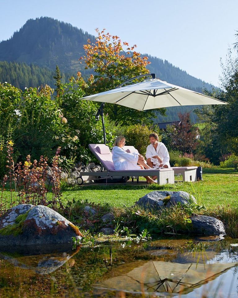 Zwei Personen im Bademantel auf Liegen entspannen sich unter einem Sonnenschirm im Garten neben Teich und Bergen im Hintergrund.