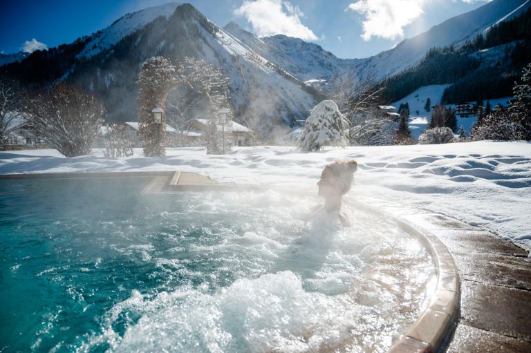 Person entspannt im dampfenden Außenwhirlpool, umgeben von tief verschneiten Bergen und klarem Himmel.