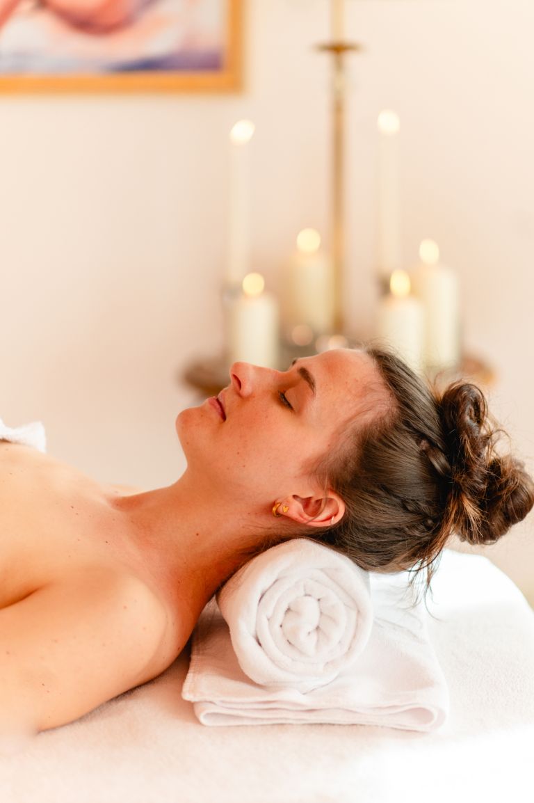 Frau liegt entspannt mit geschlossenen Augen auf einem Massagetisch mit dem Kopf auf gerolltem Handtuch und Kerzen im Hintergrund.