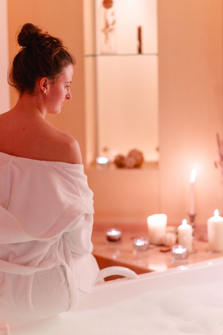 Een jonge vrouw in een witte badjas zit met opgestoken haar aan de rand van een bad, omringd door brandende kaarsen in een sfeervol verlichte wellnessruimte