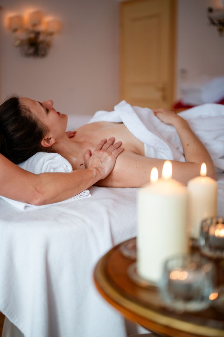 Person liegt auf Massagebank und erhält Schulter- und Brustmassage; Kerzen flackern im Vordergrund.