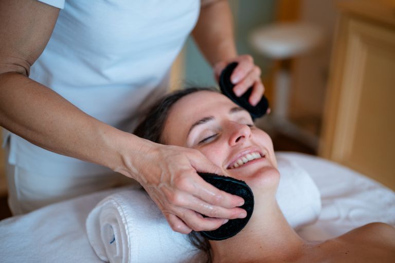 Frau lächelt während sie eine Gesichtsbehandlung mit schwarzen Pads erhält und liegt entspannt auf einem Handtuch im Spa.