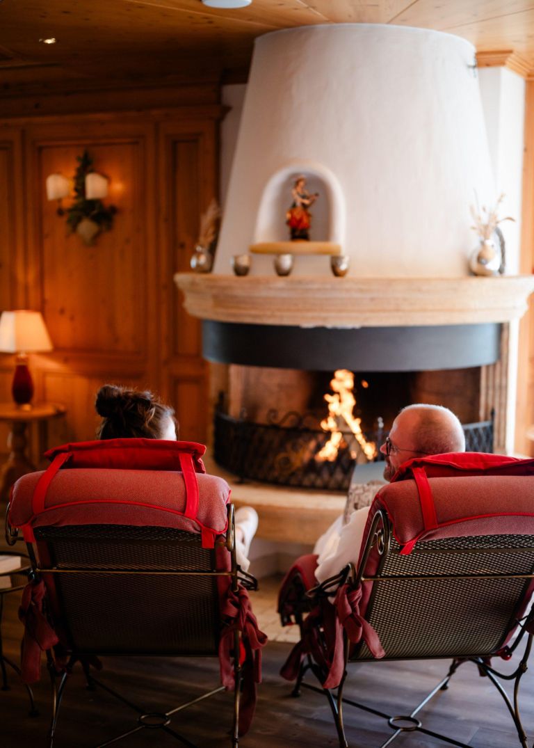 Zwei Personen sitzen in roten Liegestühlen vor einem brennenden Kamin in einem warmen, holzgetäfelten Wohnzimmer.
