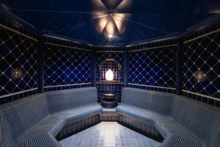 Blau gefliester Dampfraum: geschwungene Mosaiksitzbank umgibt eine beleuchtete Nische mit Salzkristall und kleinem Brunnen.