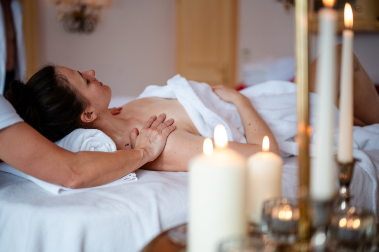 Frau liegt entspannt auf dem Rücken auf einer Massageliege, Therapeut massiert ihre Schultern, brennende Kerzen im Vordergrund.