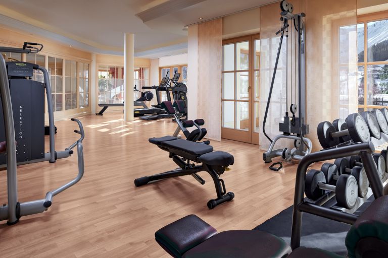 Hantelbank steht bereit, umgeben von Trainingsgeräten in hellem Fitnessraum mit Holzboden und Fenstern.