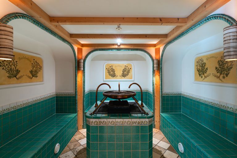 Runde Bronzeschale steht auf gefliestem Podest in einem grünen Kachelraum mit Nischenbänken und Kräuterbildern.