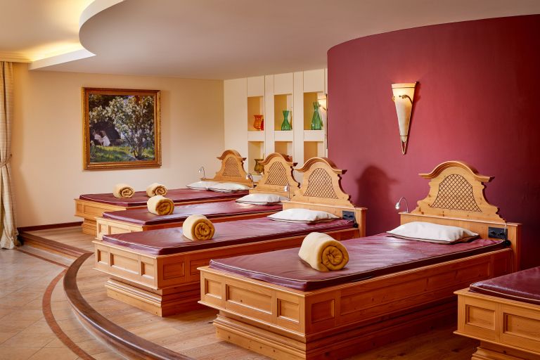 Fünf Holzliegen nebeneinander, jeweils mit zusammengerolltem Handtuch in ruhigem Wellnessraum mit roter Wand und Gemälde.