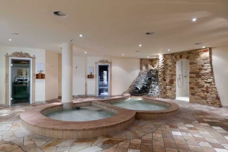 Zwei runde Thermalbecken liegen mit klarem Wasser in einem hellen Wellnessraum vor Natursteinwand, kleinem Wasserfall und Dusche.