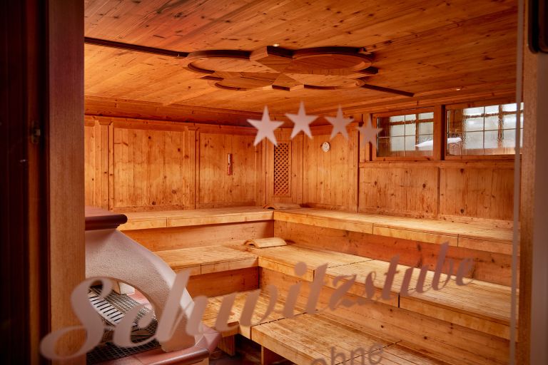 Holzsauna mit gestaffelten Sitzbänken und Ofen durch ein Fenster mit der Aufschrift "★★★★ Schwitzstube" zu sehen.