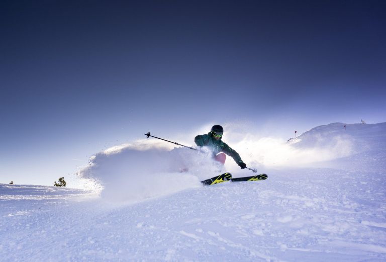 Skifahrer schneidet in Pulverschnee, wirbelt eine Spraywolke auf, auf einem breiten, sonnigen, verschneiten Hang.