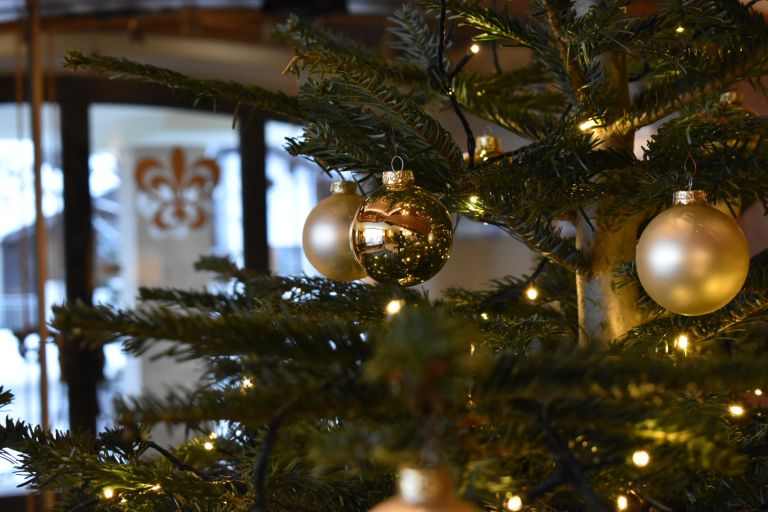 Goldene Weihnachtskugeln hängen an einem beleuchteten Tannenbaum im warmen Innenraum vor einem unscharfen Fenster.