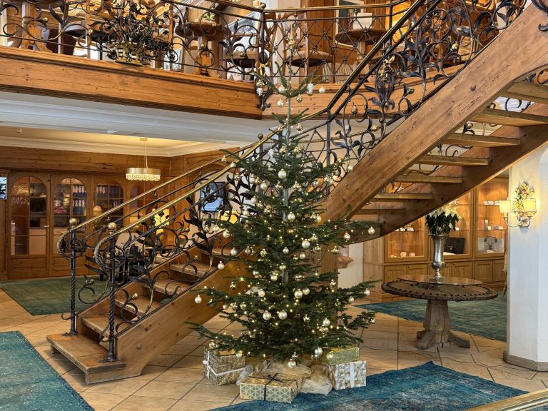 Weihnachtsbaum, geschmückt mit Lichterketten und goldenen Kugeln, steht vor geschwungener Holztreppe in eleganter Hotellobby.