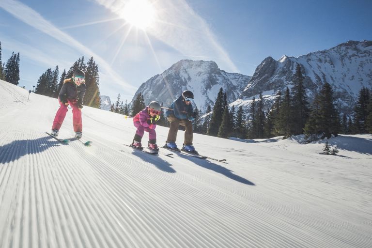Drei Skifahrer in leichter Hocke fahren die frisch präparierte Piste hinunter, vor sonnigen, schneebedeckten Bergen.