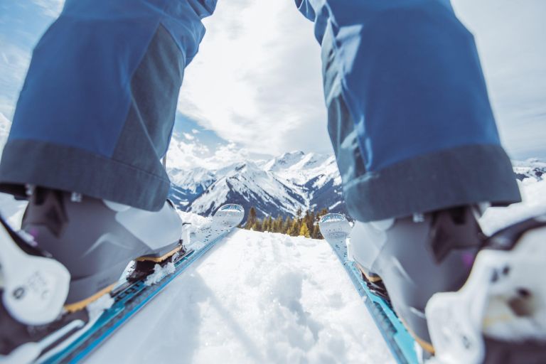 Skischuhe in Bindungen stehen auf Skiern, bereit zu fahren, vor verschneiten Alpenbergen unter blauem Himmel.