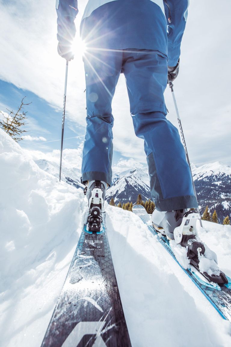 Skifahrer steht auf Skiern und hält Stöcke; Sonnengeflutete, verschneite Berglandschaft im Hintergrund.