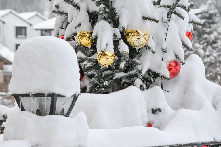 Weihnachtsbaum mit goldenen und roten Kugeln, Zweige schwer vom Schnee bedeckt, im verschneiten Außenbereich vor Häusern.