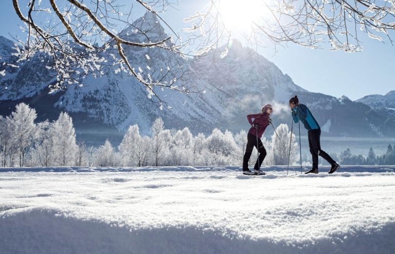 Zwei Skilangläufer lehnen an Stöcken und unterhalten sich auf verschneiter Ebene vor frostigen Bäumen und Bergen bei Sonnenschein.