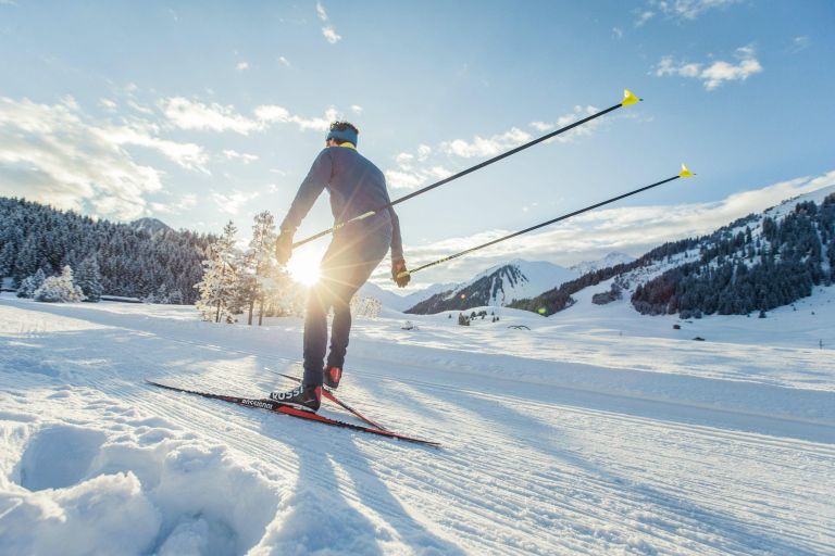Langläufer gleitet über gespurte Piste, Sonne blitzt zwischen verschneiten Bergen und Nadelwald hervor.
